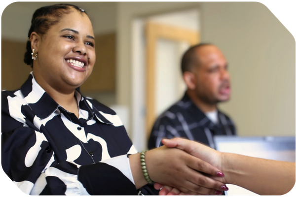 black woman in black and white patterned blouse shaking hands with an unseen recruiter. A black man is being interviewed in the background