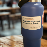 A water bottle on a cafe table with a branded sticker saying: Say more about that. What mess do you need permission to make?