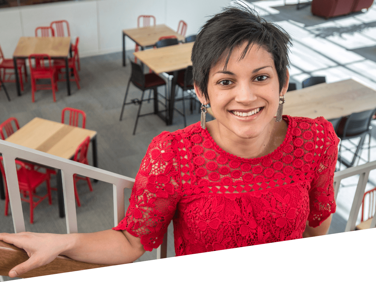 Brown-skinned woman with short brown hair wearing a red lace top standing on a staircase with cafe tables behind her Brown-skinned woman with short brown hair wearing a red lace top standing on a staircase with cafe tables behind her